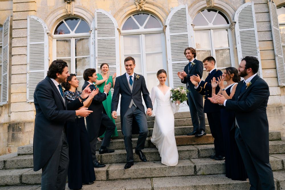 Wedding party celebrates on stone steps outside historic building while taking group photos.