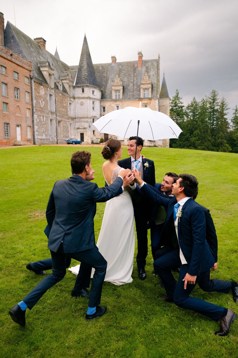 Four groomsmen in navy suits playfully pose on grass lawn outside historic chateau in France.
