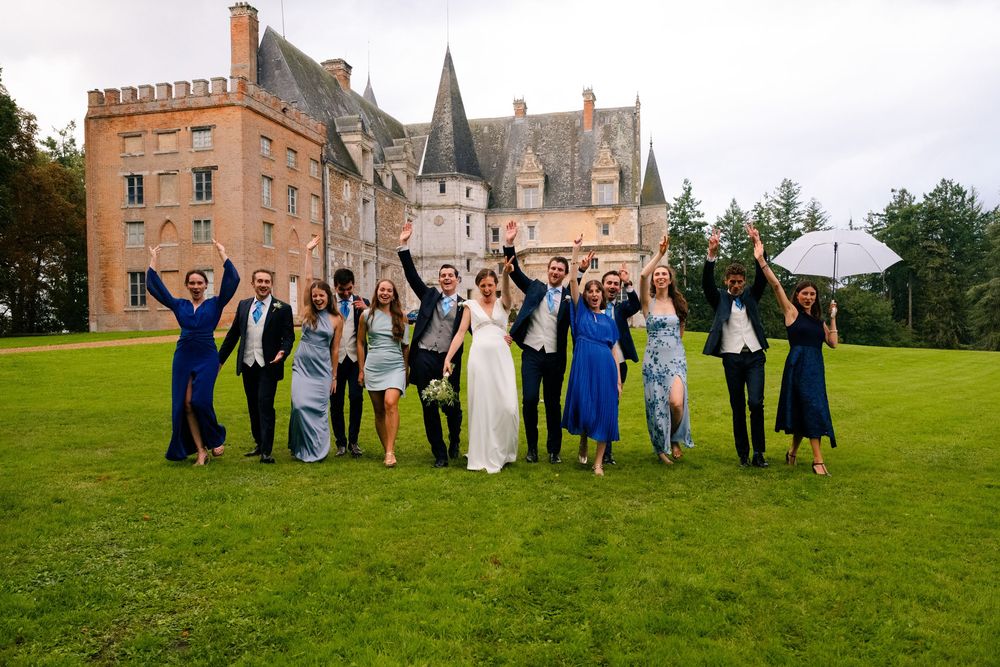 Wedding party members jump joyfully on the green lawn outside a historic brick castle building during celebration.
