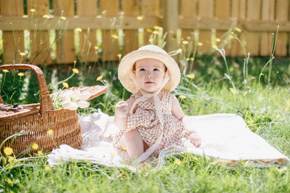 Baby in polka dot outfit and sun hat sits on blanket during backyard picnic.