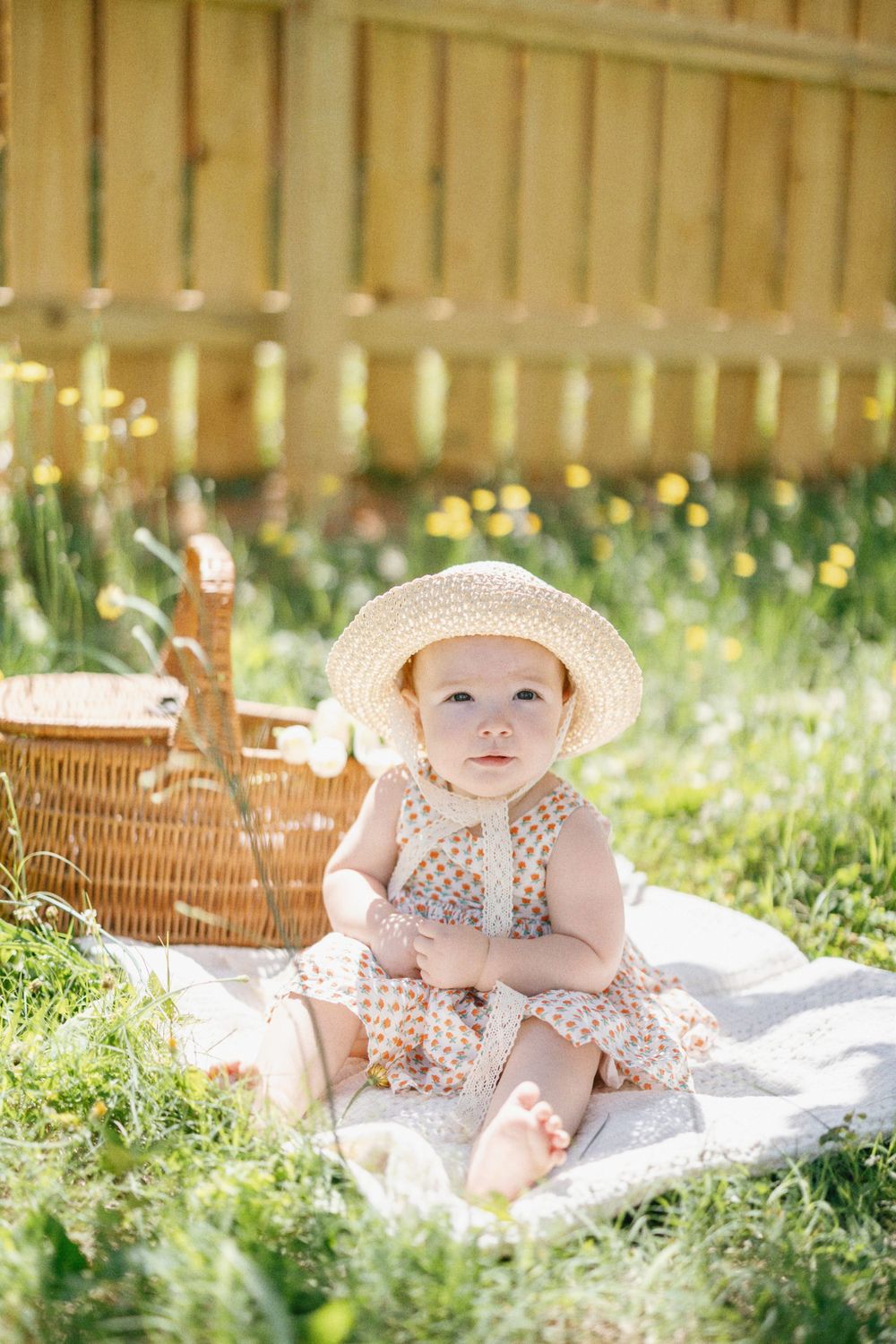 Baby in summer dress and sun hat sits on blanket by picnic basket in sunny garden.