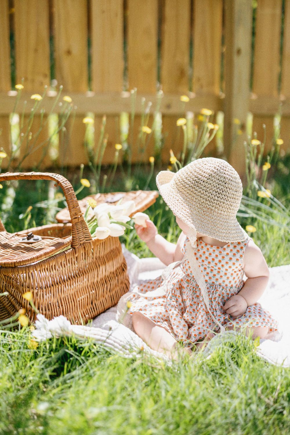 Child in spotted dress reaches for picnic basket while sitting on blanket outdoors.