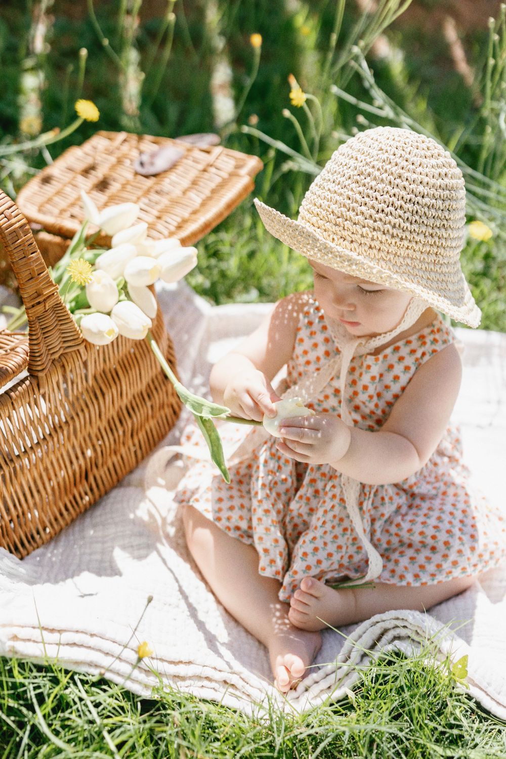 Baby in spotted dress examines flowers next to wicker picnic basket on blanket.