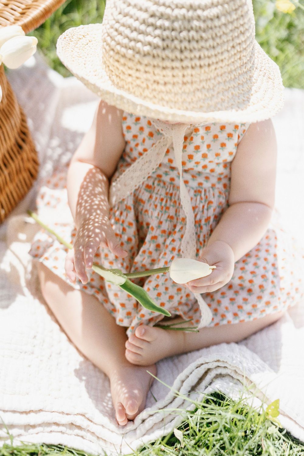 Small child in sun hat holding flower while sitting on picnic blanket.