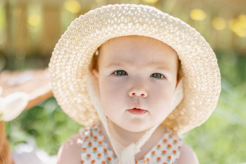 Small child wearing a floral dress and straw sun hat sits in sunny garden setting.