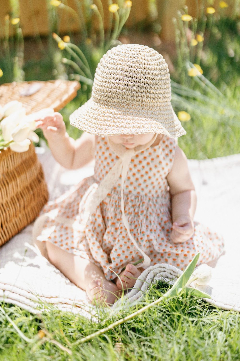 Toddler in summer dress and woven hat sits on picnic blanket near wicker basket.