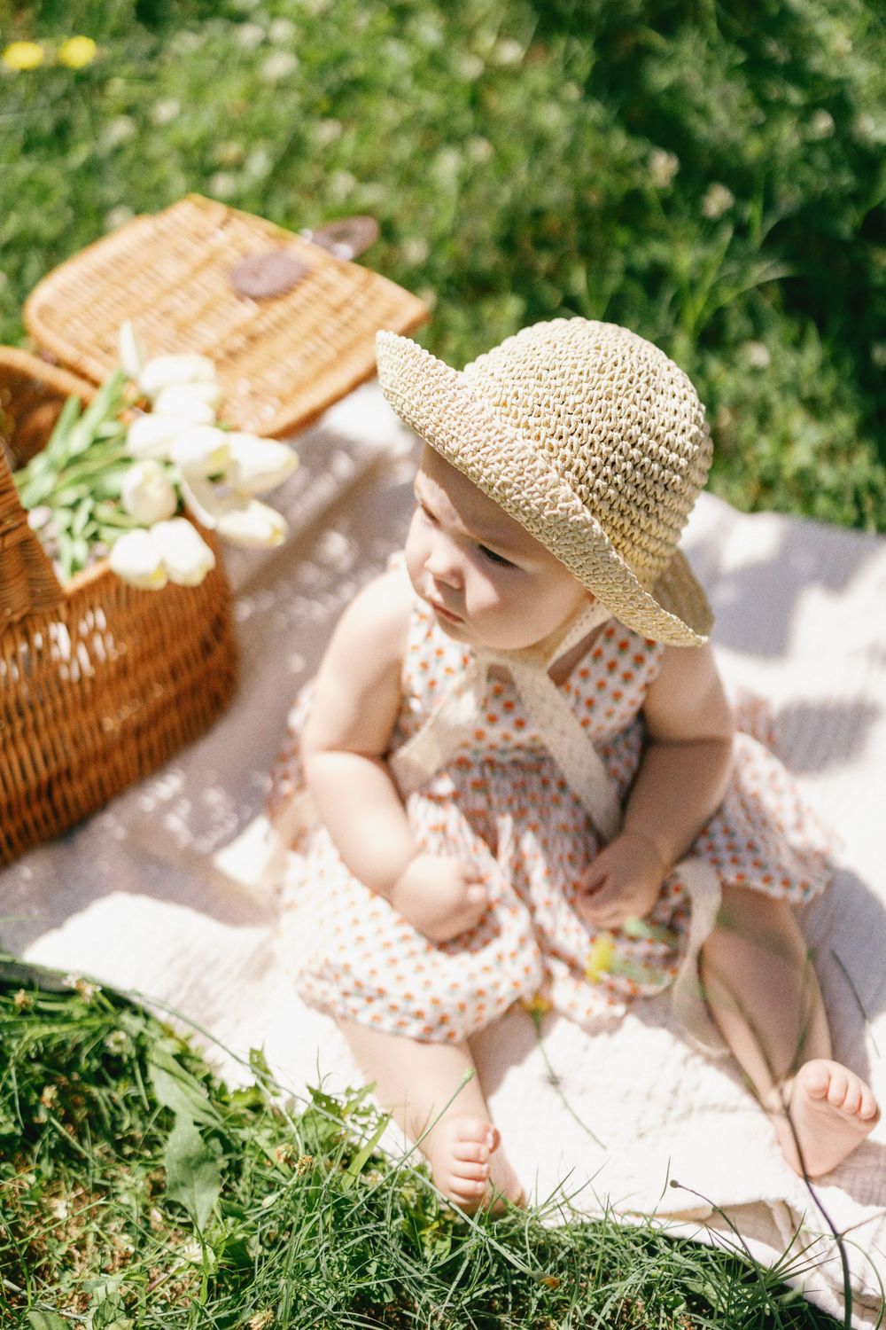 Child in spotted dress and straw hat explores during outdoor picnic on sunny day.
