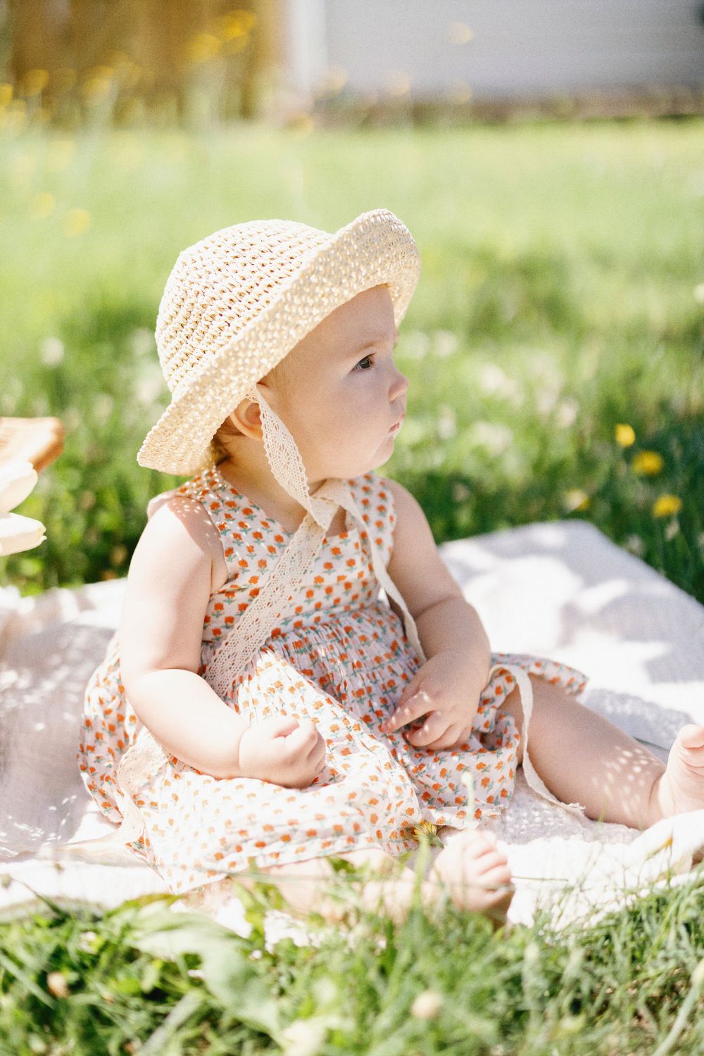 Toddler in summer dress and straw hat sits on white blanket in sunny garden.