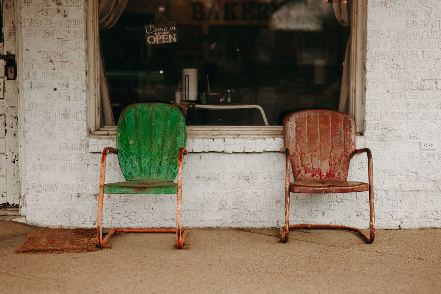 Two vintage rusty metal chairs, green and red, sit outside an old brick storefront.