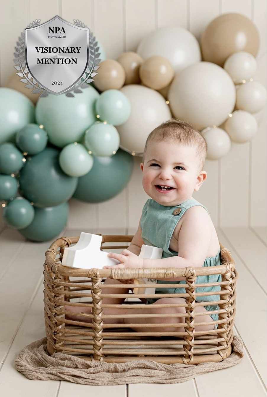 Baby wearing mint green romper sits in woven basket against balloon backdrop with NPA visionary mention award.