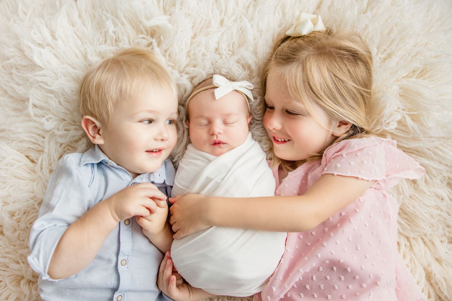 Three young siblings cuddle together on a soft white fur blanket in a sweet and tender moment.  Middle TN Newborn Photographer.