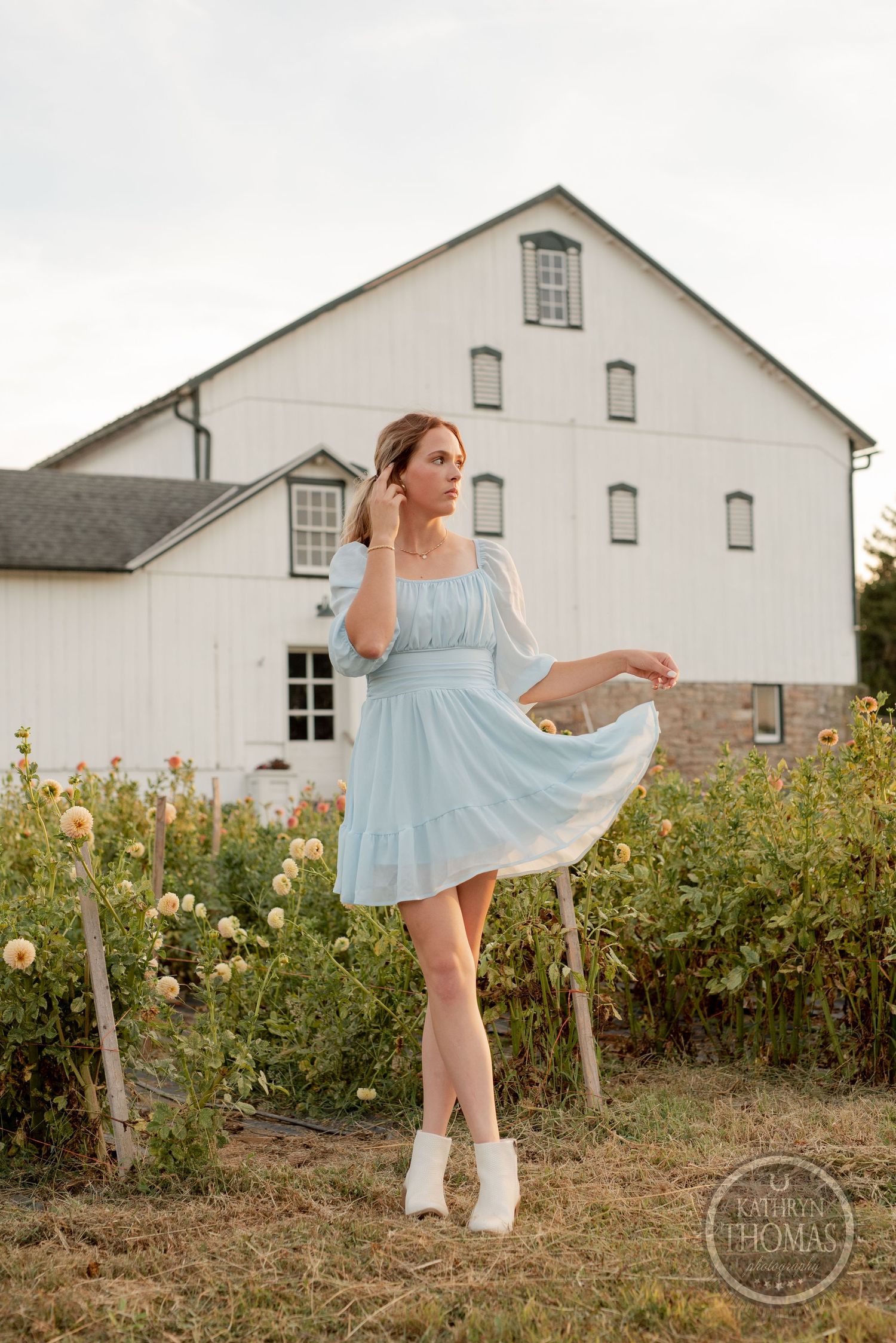 A graceful pose in a light blue dress against a white barn with orange flowers in the foreground.