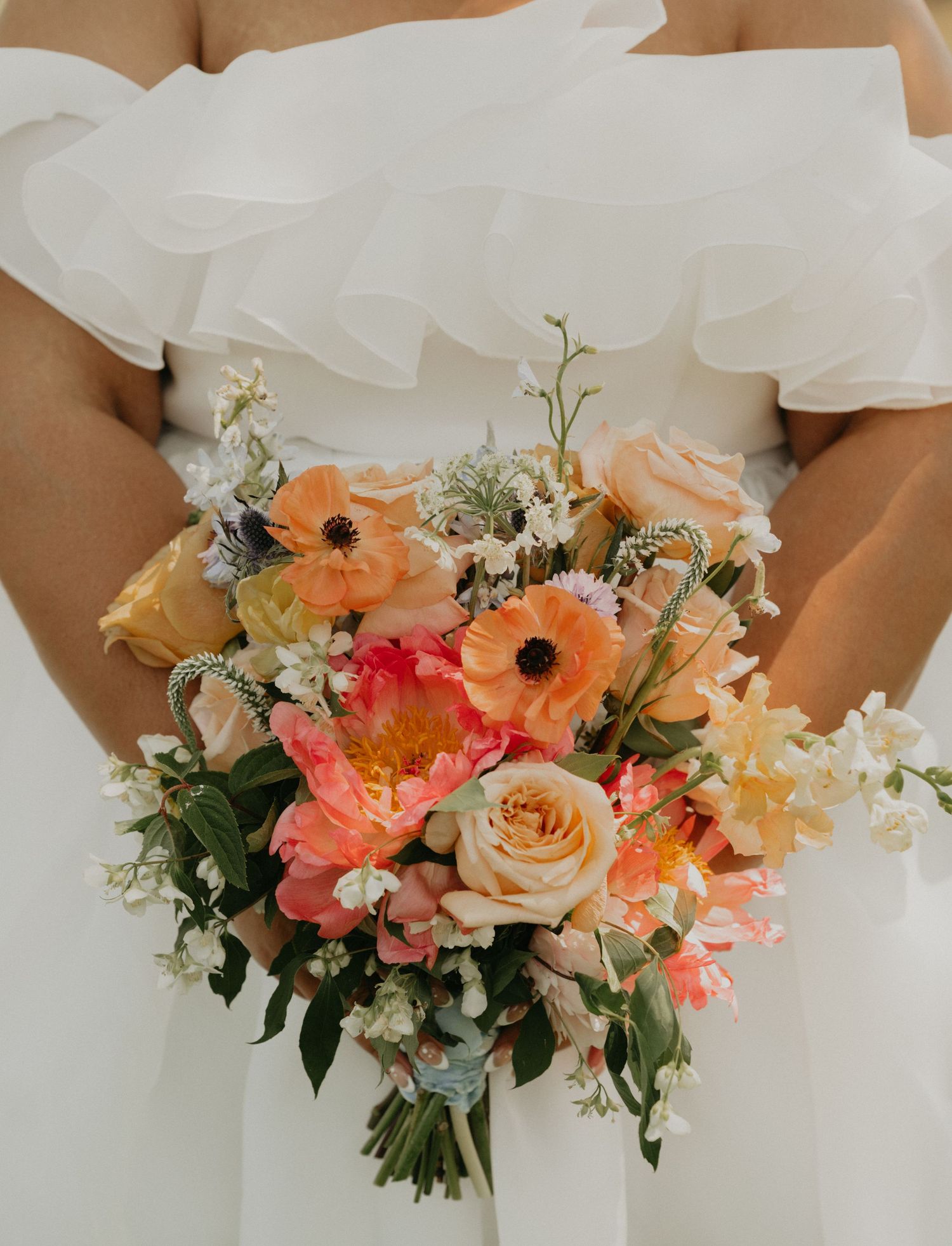 Bride holding vibrant coral and peach wedding bouquet against white dress.