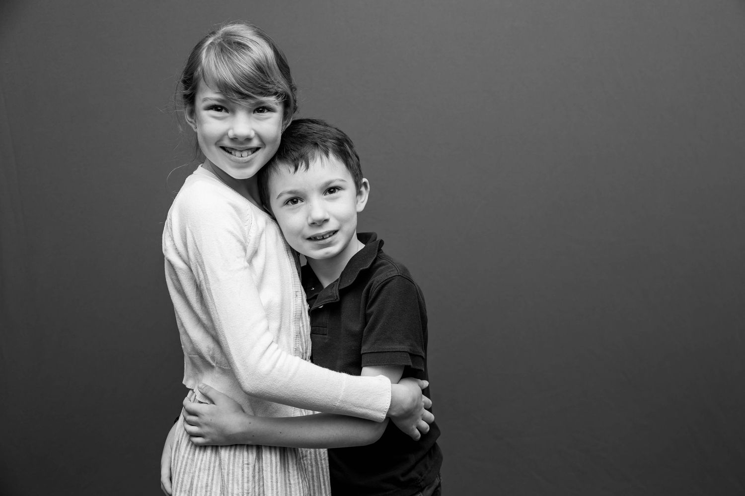Black and white portrait of two children embracing and smiling against a dark backdrop.