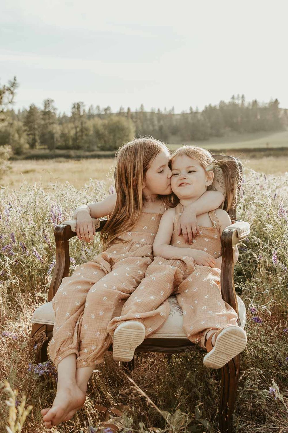 sisters sitting on a vintage chair in open flower field at golden hour with a spokane family photographer