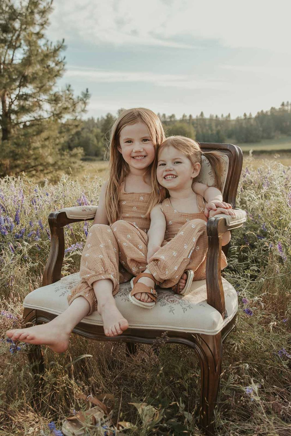 sisters sitting on a vintage chair smiling in open flower field at golden hour with a spokane family photographer