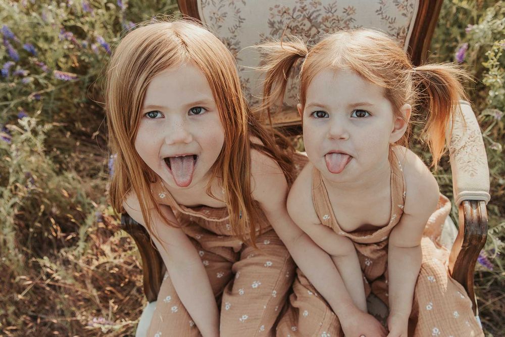 sisters sitting on a vintage chair smiling in open flower field at golden hour with a spokane family photographer