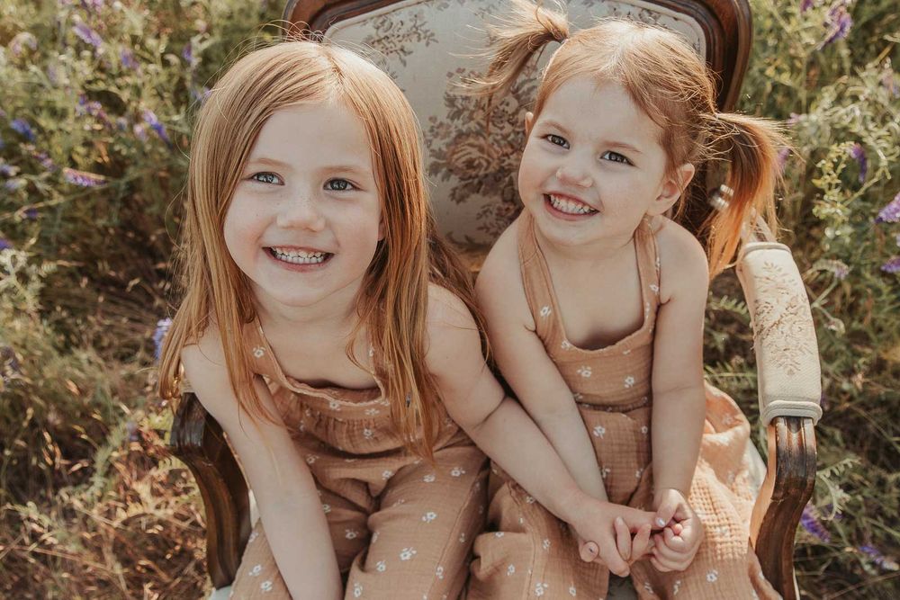 sisters sitting on a vintage chair smiling in open flower field at golden hour with a spokane family photographer