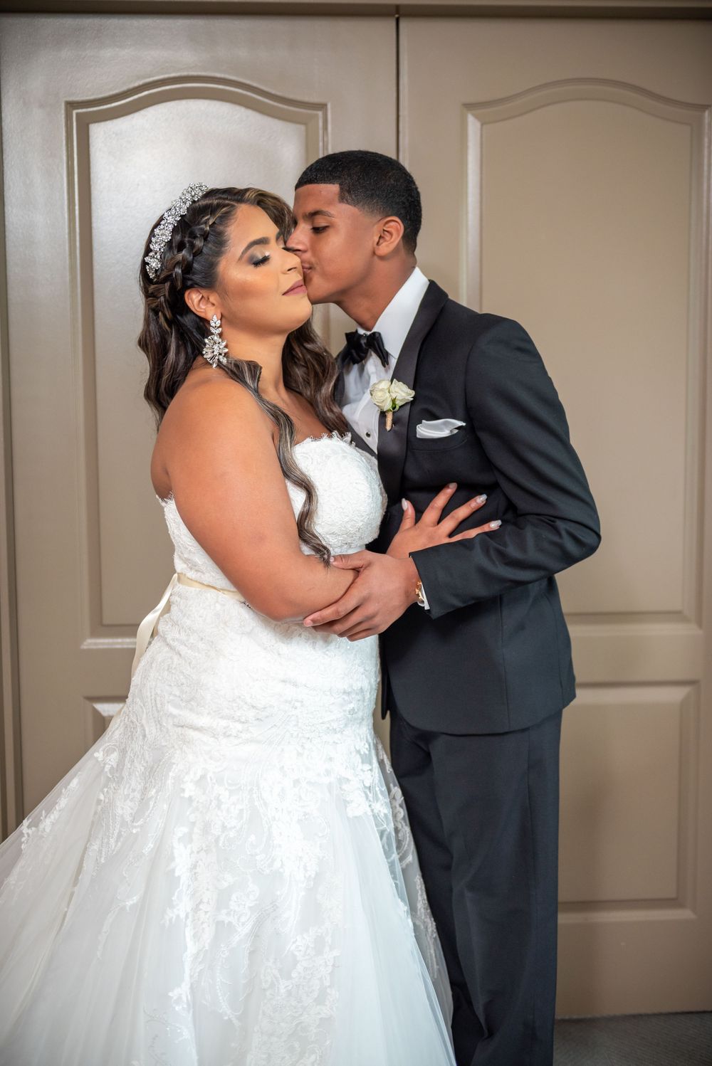 A romantic moment between newlyweds as they share their first kiss in formal wedding attire against neutral doors.
