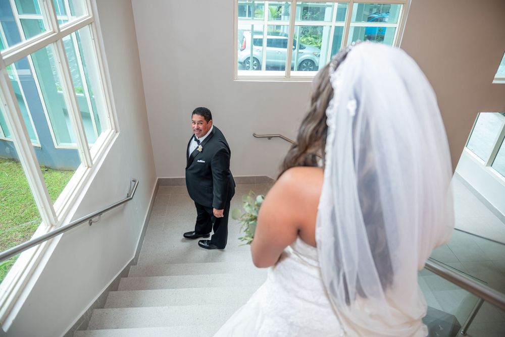 A bride in white dress and veil descends stairs while looking at someone waiting below.