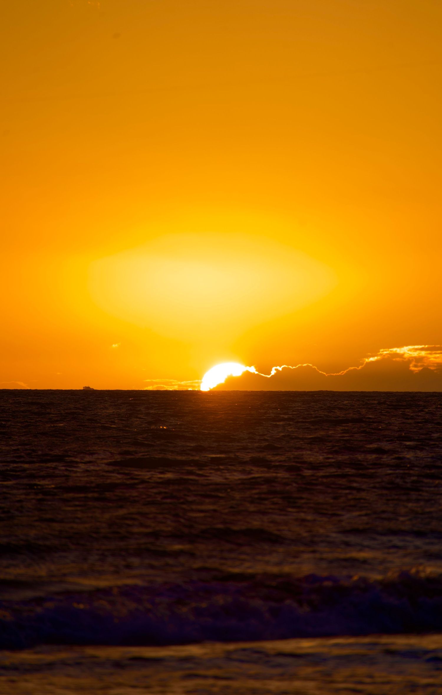 Dramatic orange sunset over dark ocean waters with sun dipping below cloud-lined horizon.