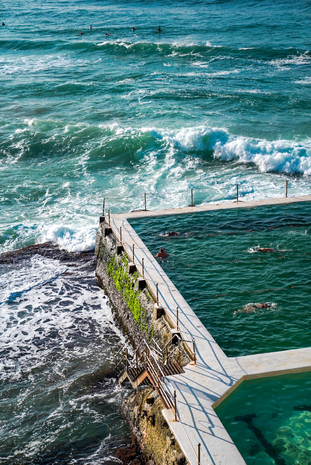 Ocean pool with concrete barrier wall separating calm turquoise waters from crashing waves.