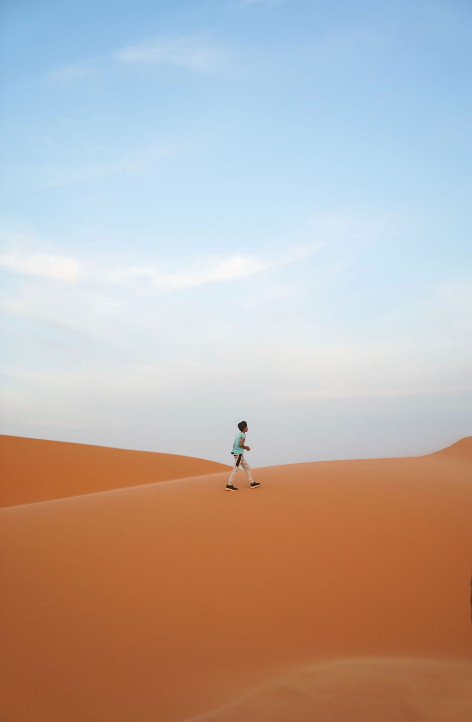 A lone figure walks across rippling orange sand dunes under a pale blue desert sky.