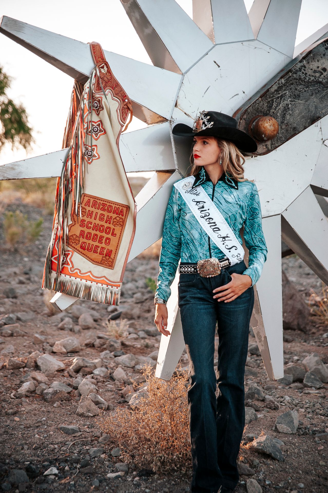 Rodeo queen poses against rustic white star backdrop in desert setting.