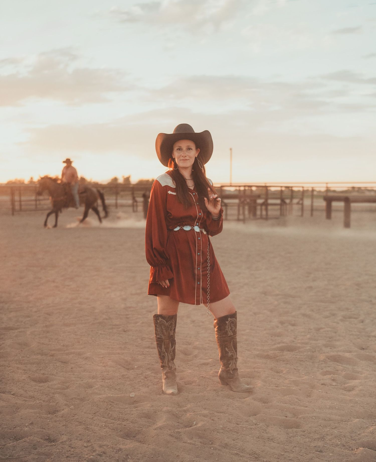 Cowgirl in a red dress and cowboy hat sits in a grassy field at sunset next to a grazing horse.
