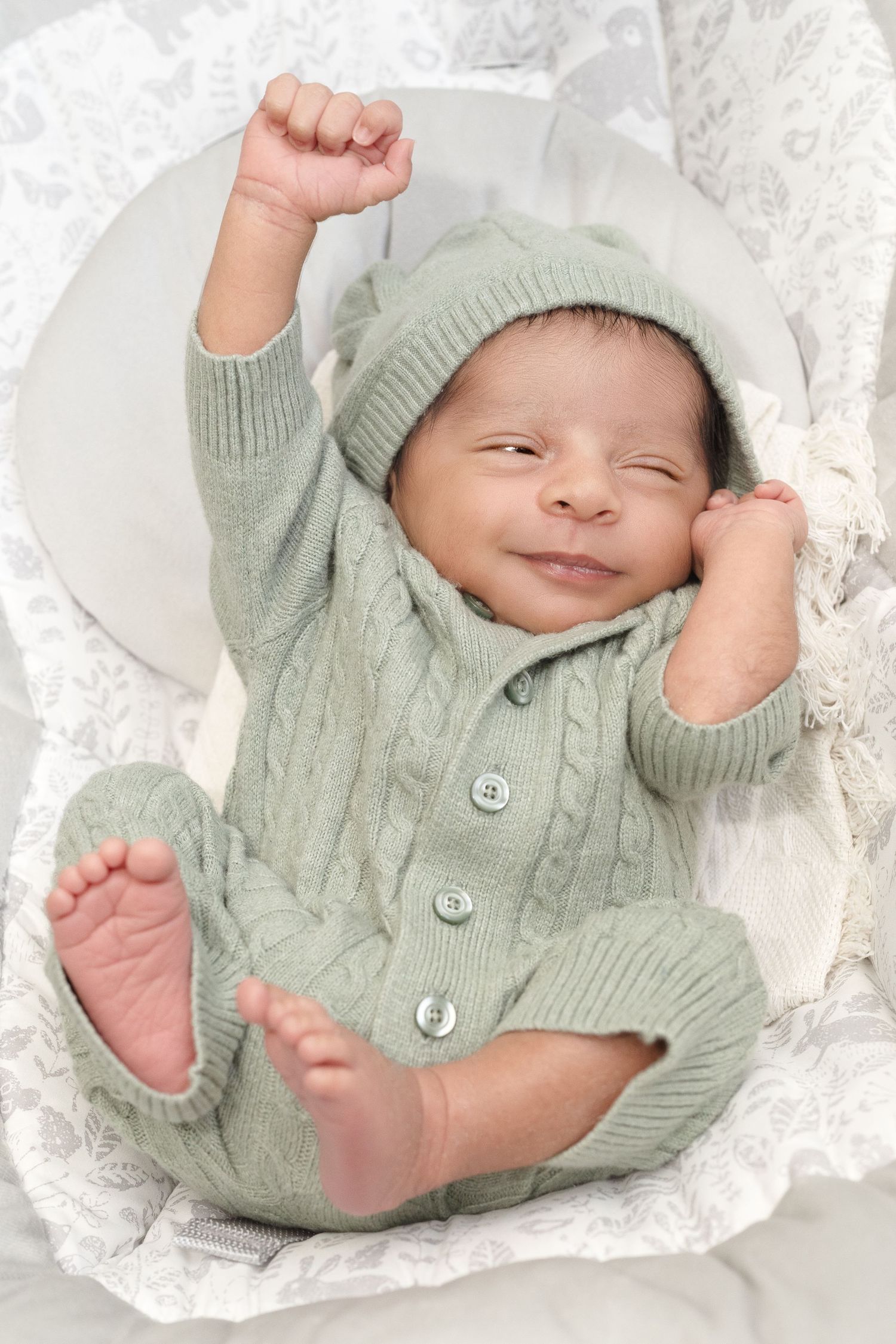 A newborn baby wearing a mint green knitted sweater and beanie stretches happily on white blankets.