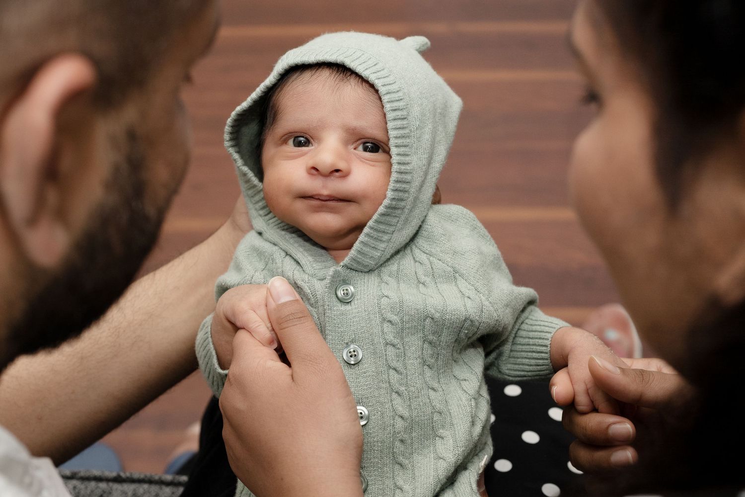 A baby wearing a sage green knitted outfit smiles while being held.