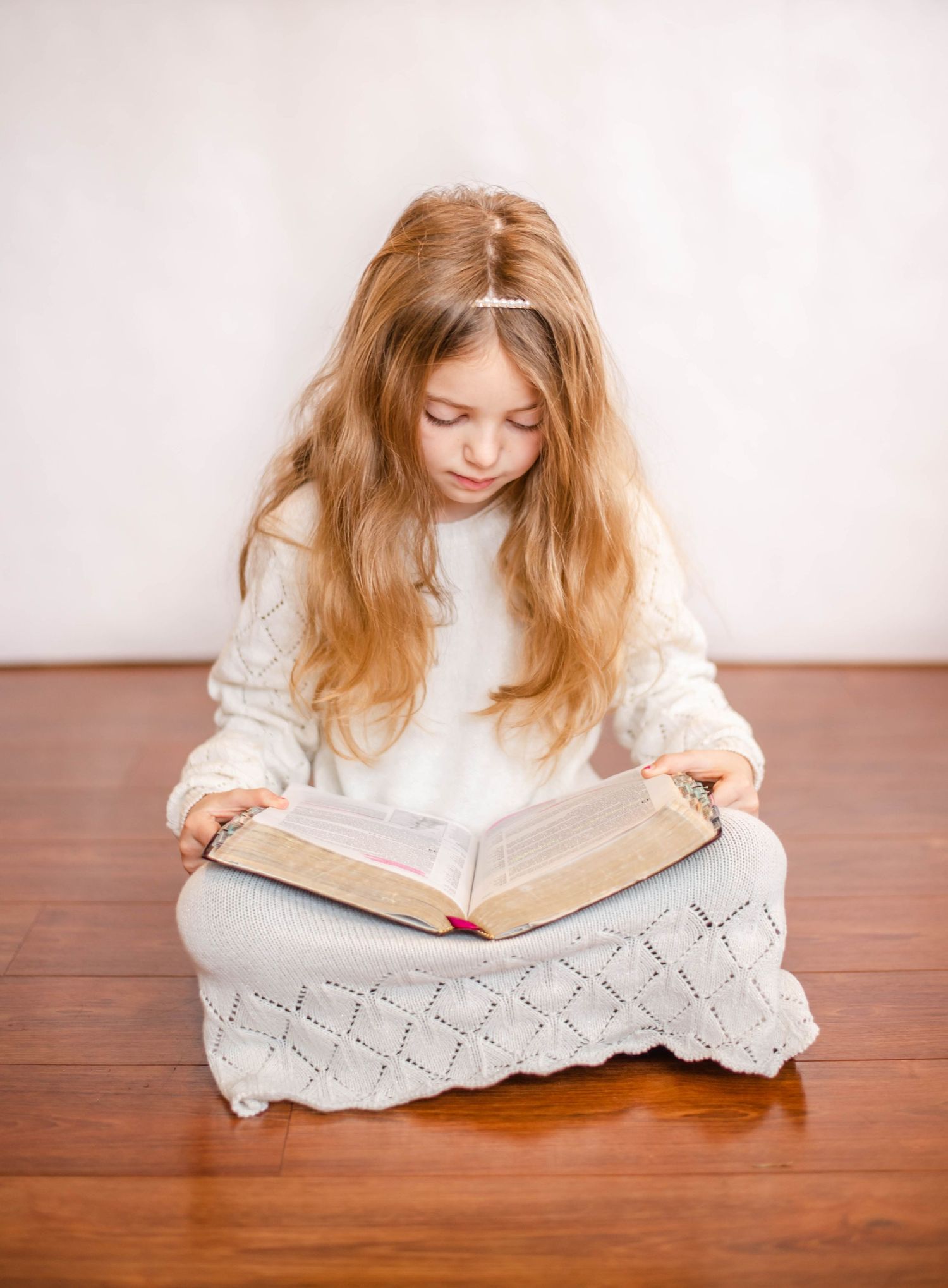 Person in white sweater reads intently from book while sitting on gray knit cushion.