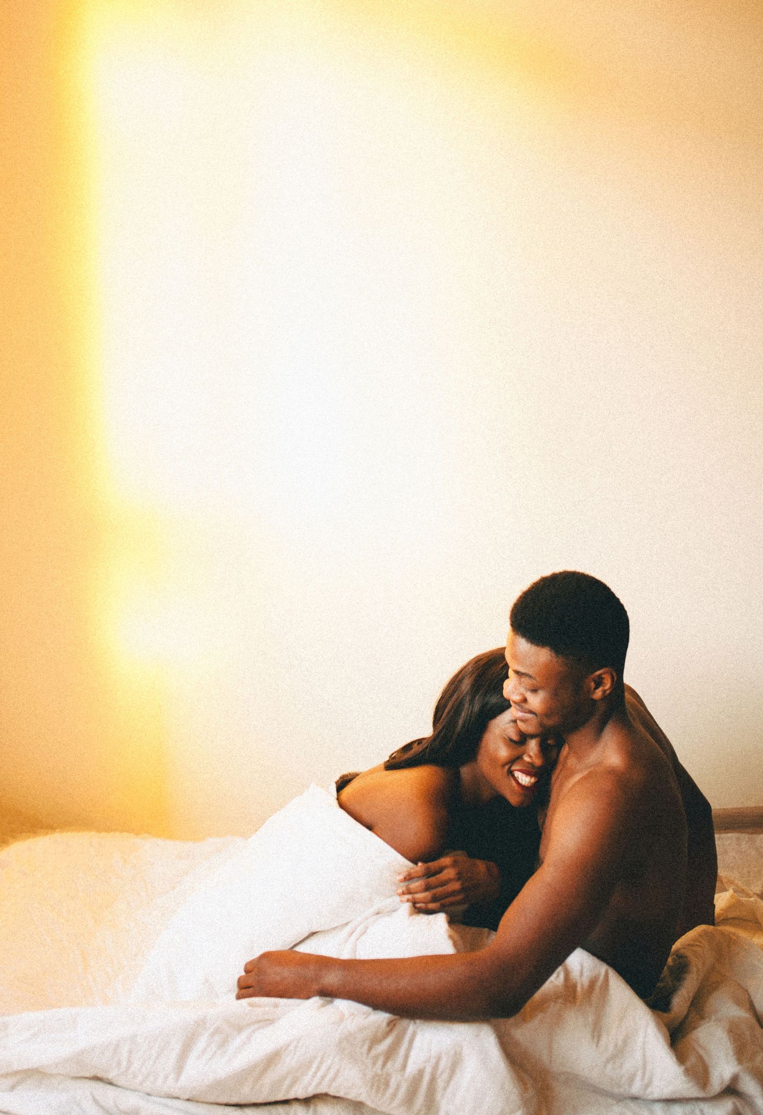 An intimate couple embraces while relaxing together on white bedding in a room with warm golden lighting.