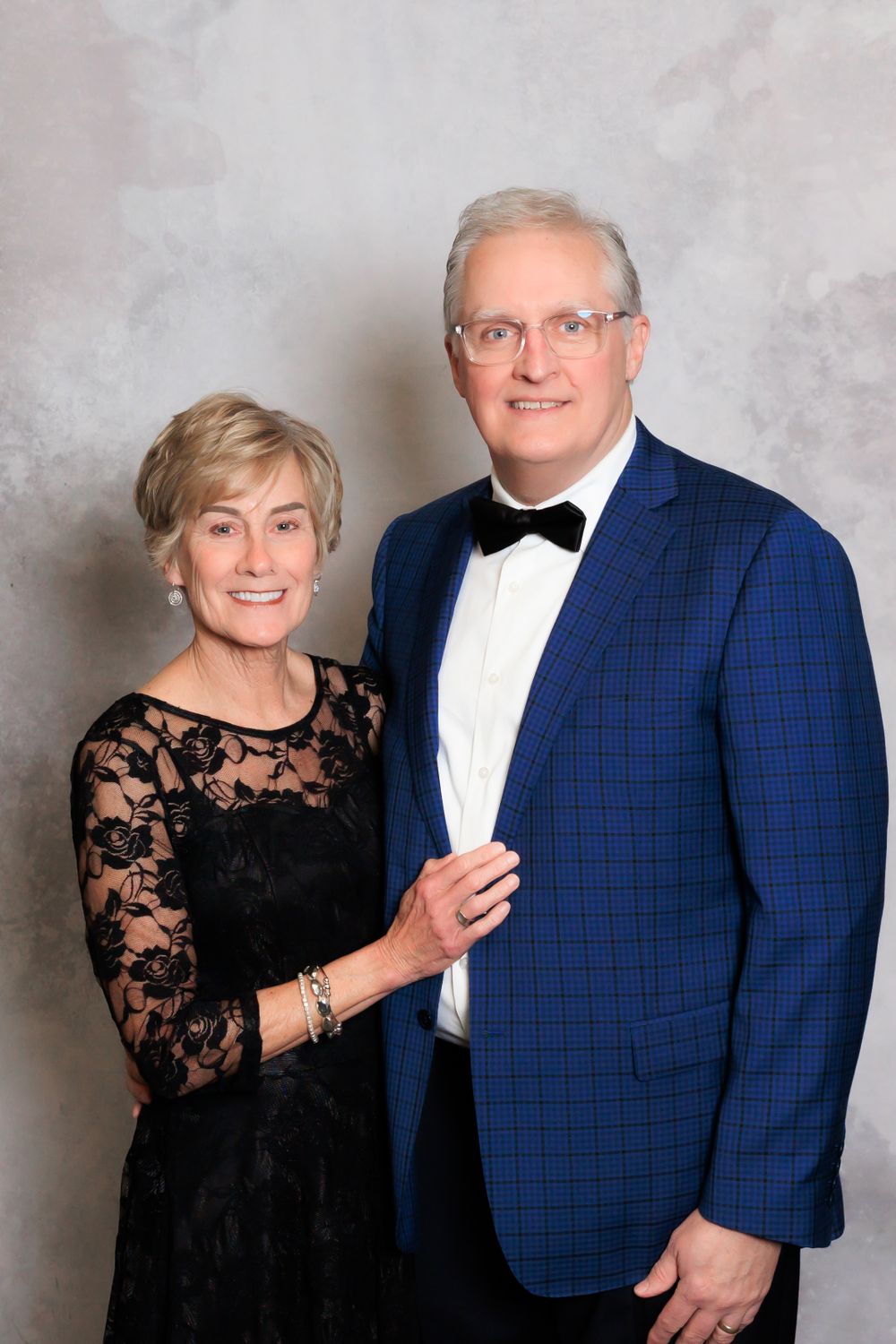 Formal couple in evening wear posing against textured grey background.
