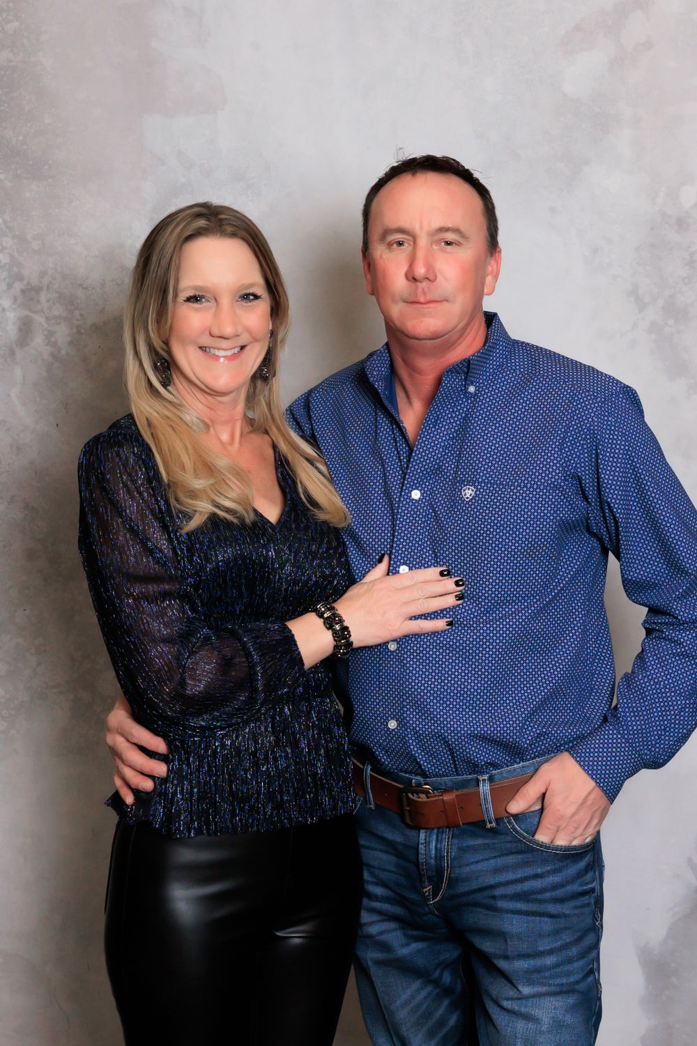 A couple poses together for a formal portrait against a gray backdrop, wearing blue and black evening attire.
