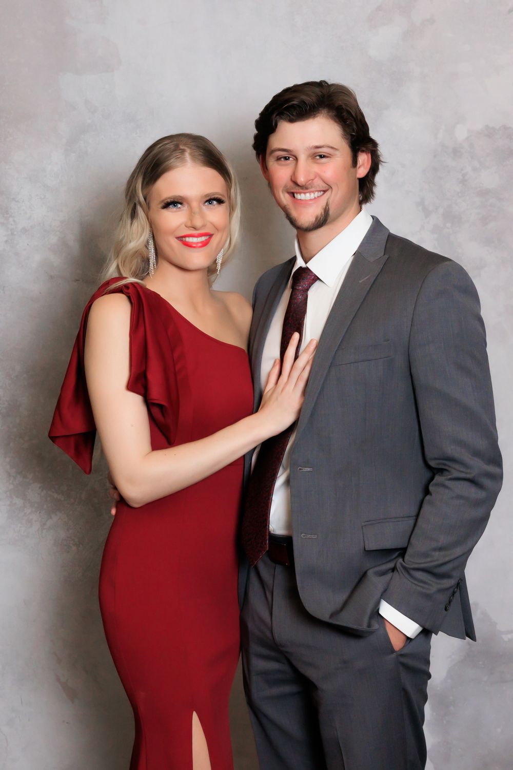 A couple poses for a formal portrait with the woman in a red gown and man in gray suit.
