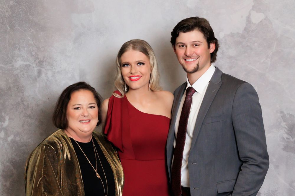 Three people pose for a formal portrait against a gray background wearing elegant evening attire.