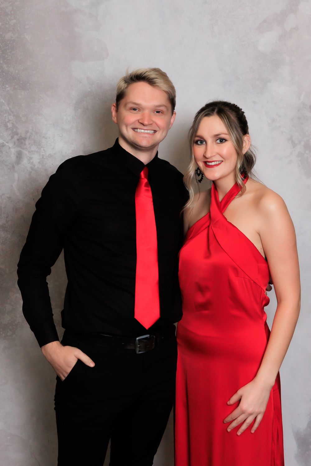 A couple poses for a portrait wearing red and black formal attire against a gray backdrop.