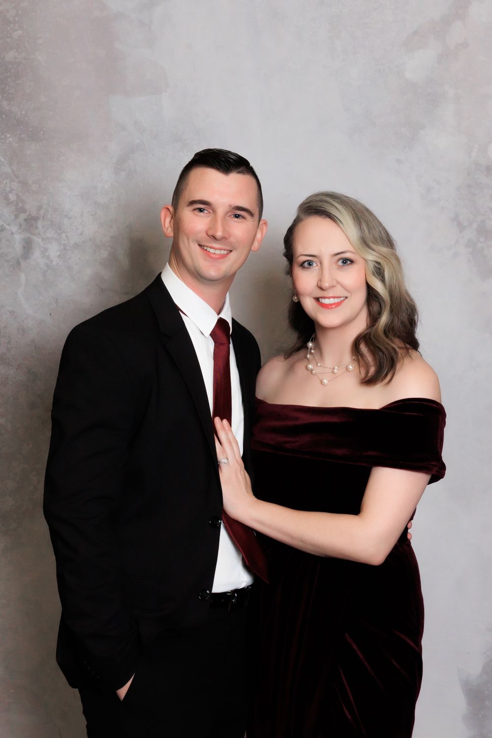 A couple poses formally against a gray background, with the woman in a burgundy velvet dress.