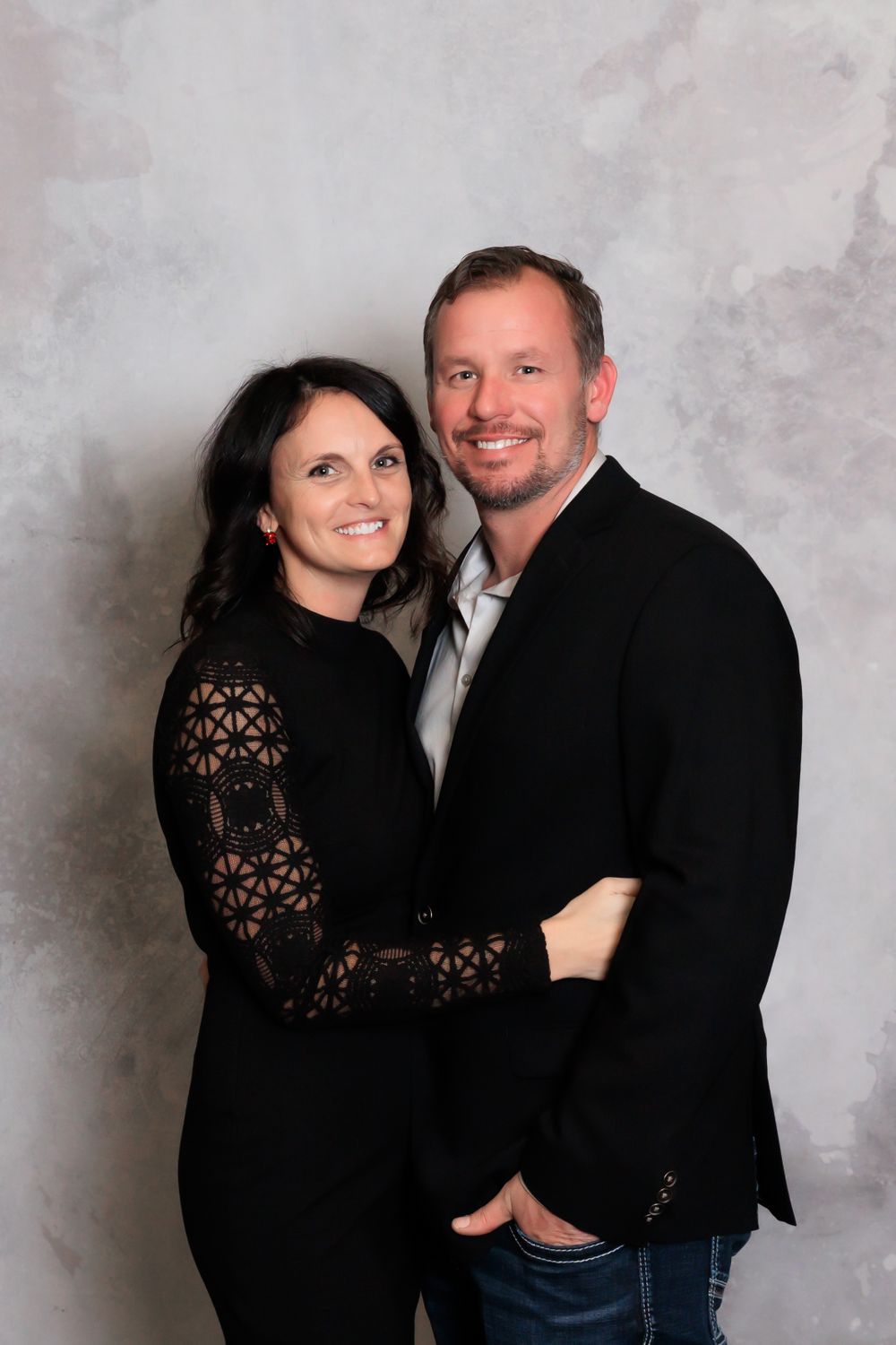 A couple poses together in black evening wear against a gray studio backdrop.