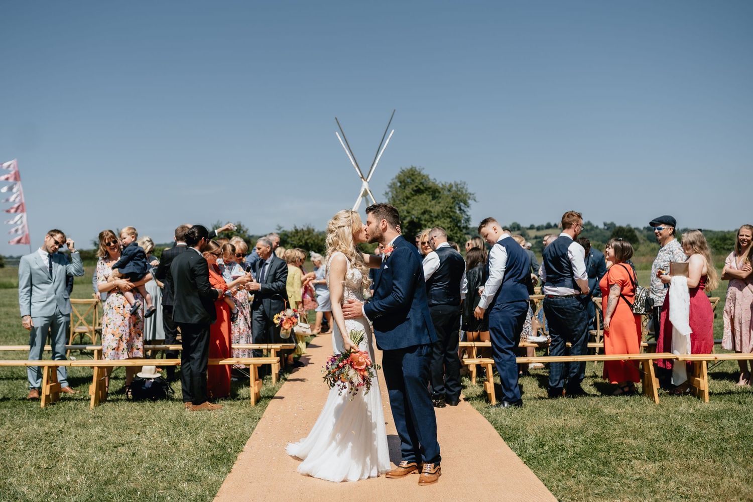 An outdoor Worcestershire wedding ceremony takes place along a tan aisle runner with guests standing on benches under clear blue skies.
