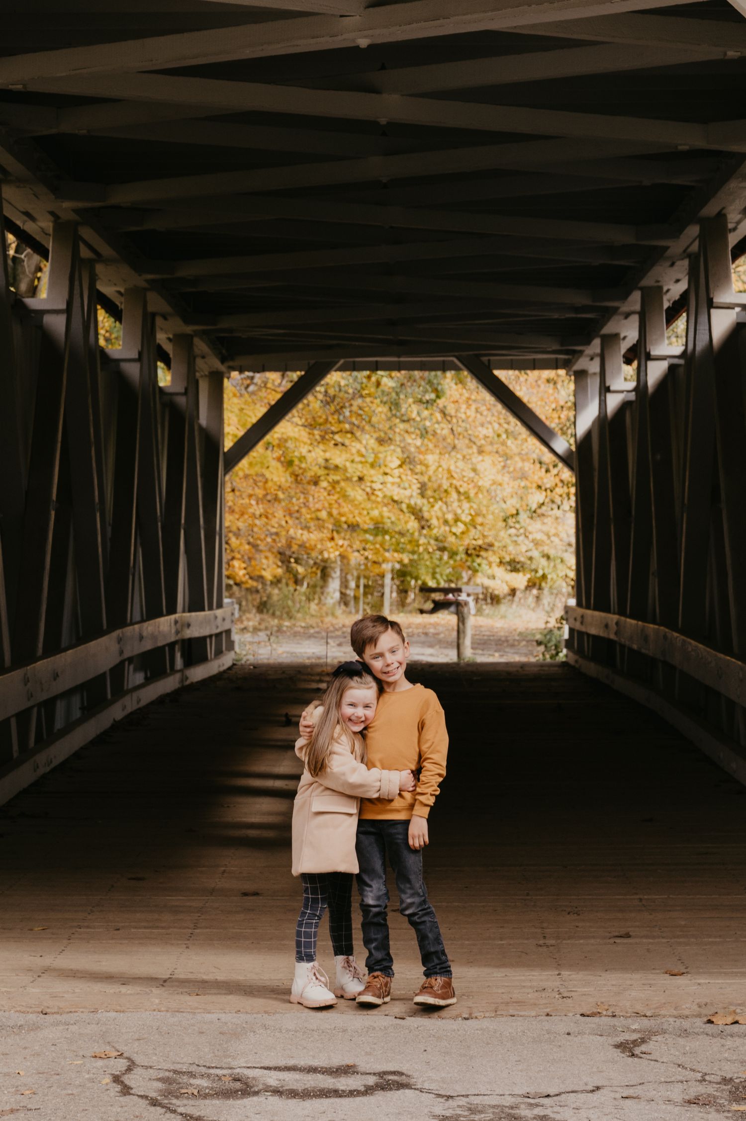 Two young siblings hugging on a covered wooden bridge surrounded by golden fall foliage.
