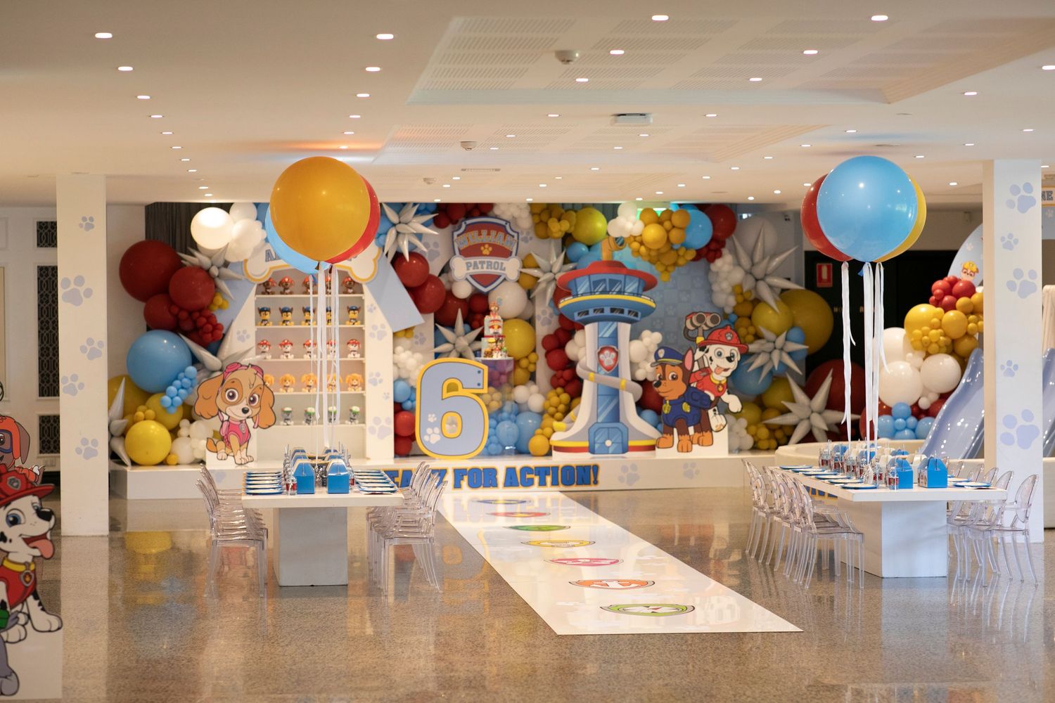 A decorated party room with colorful balloons, cartoon character wall art, and long white tables set up for a celebration.