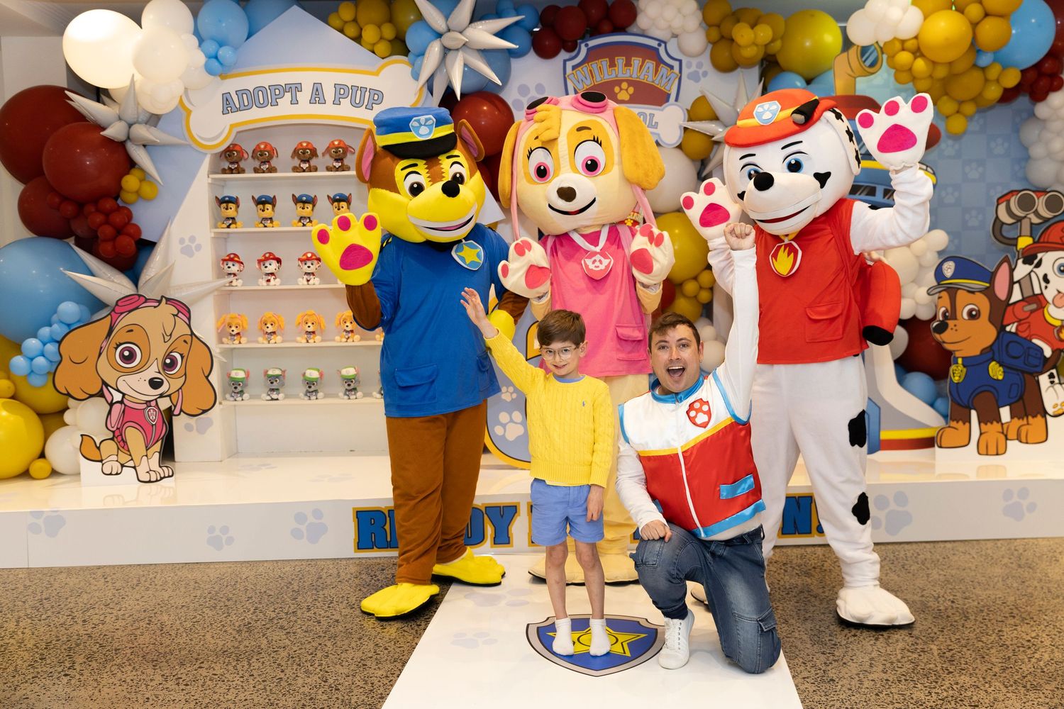 A series of mascot characters in costumes pose together at what appears to be a promotional event with colorful balloons.