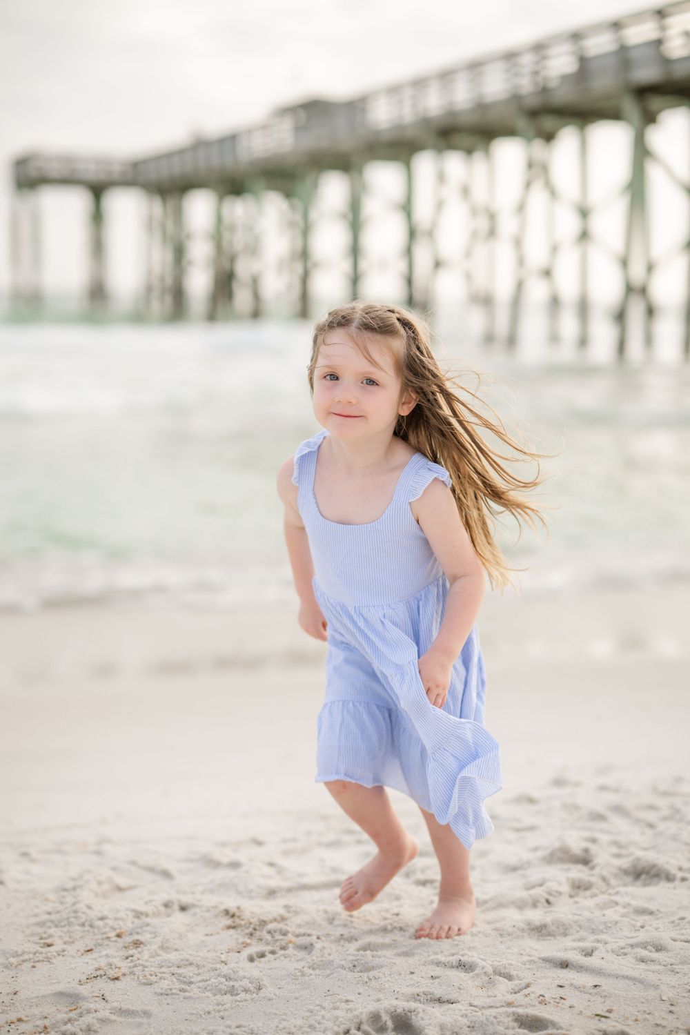 Young girl in light blue dress playing joyfully on the beach near a pier.