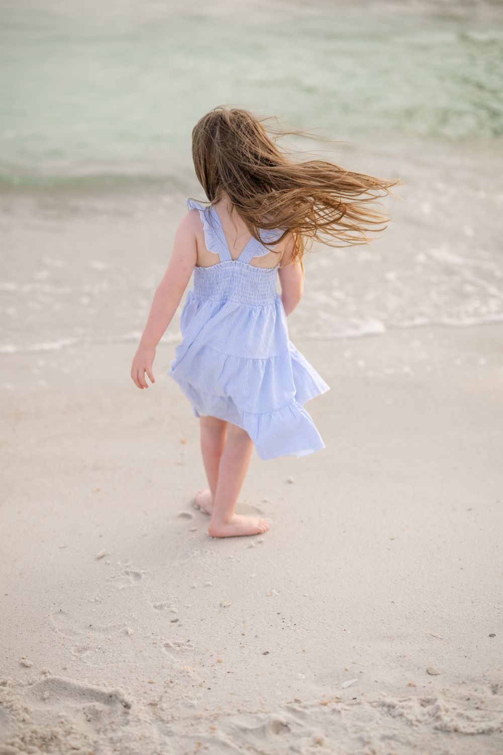 Child in flowing blue dress walking along beach with hair blowing in wind.