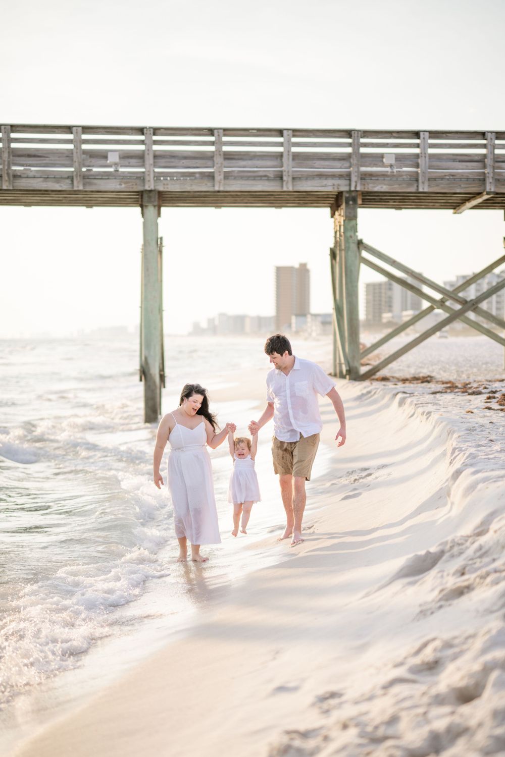 Family walking along beach shoreline under wooden pier at sunset.