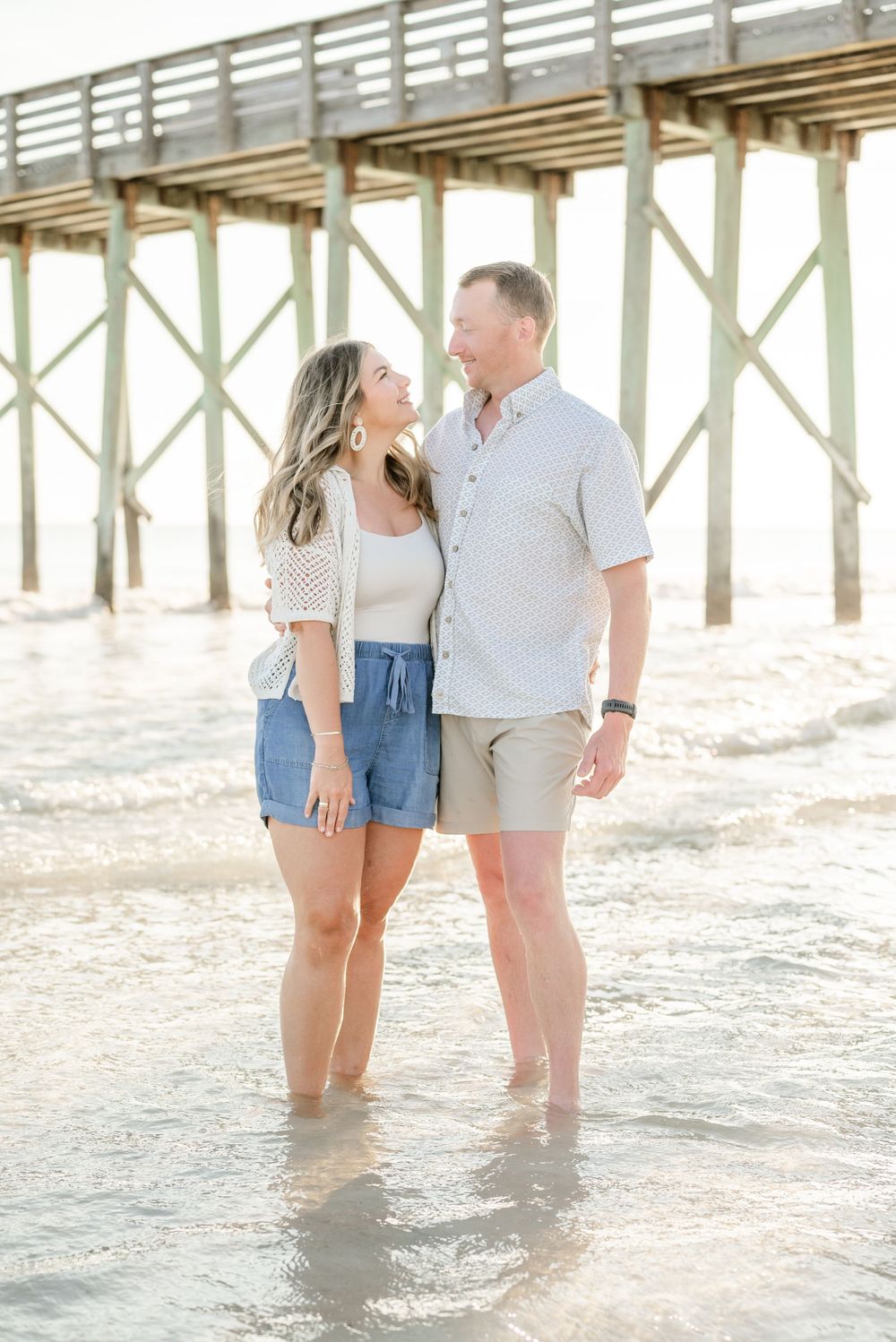 Romantic couple standing in gentle surf beneath wooden pier at beach.