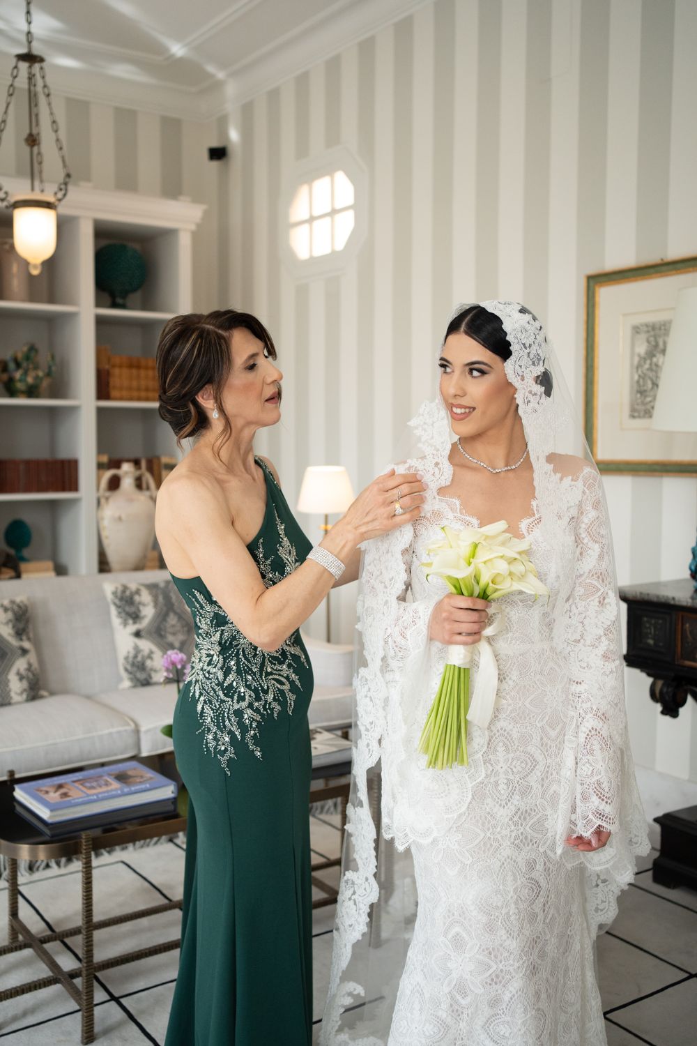 A touching pre-wedding moment between a bride in lace and attendant in emerald green dress.