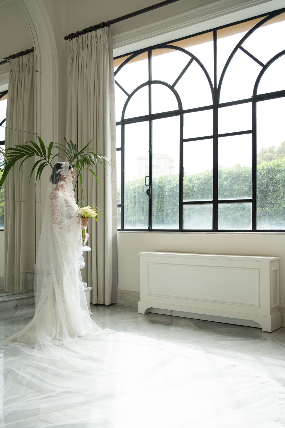 A bride in lace gown stands by arched windows in an elegant white room with palm plants.